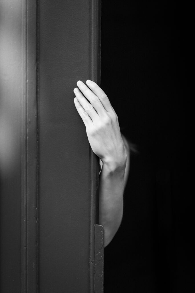 Une main et un avant-bras nu émergent de l'obscurité et se posent contre le bord clair d'une porte entrouverte, le corps invisible dans le noir, Vieille-Ville de Genève, noir et blanc.