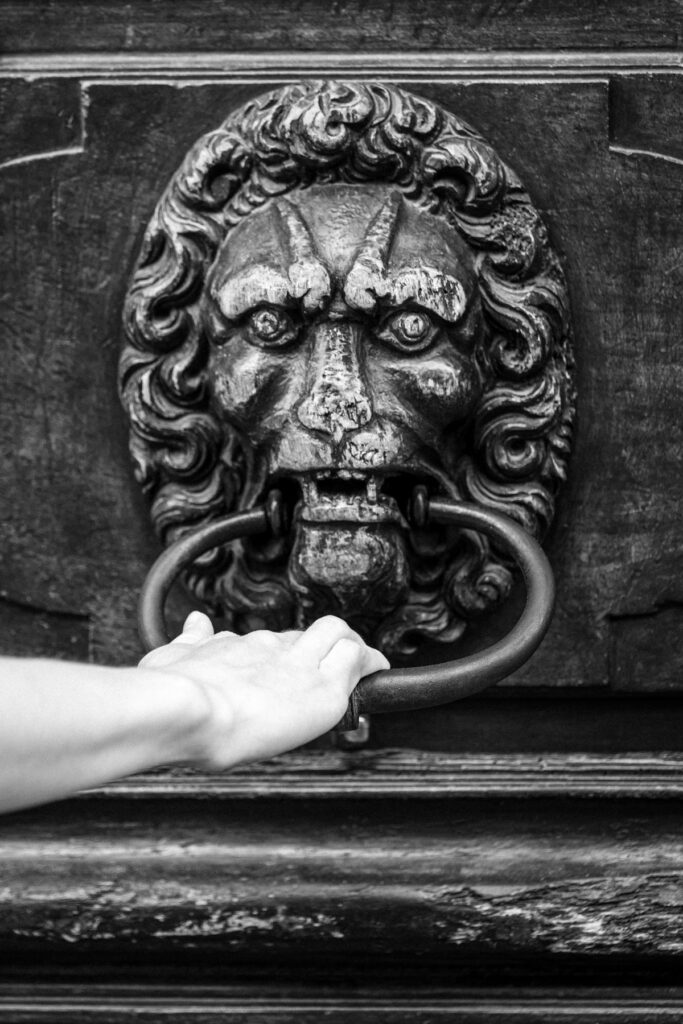 Main saisissant l'anneau de bronze d'un heurtoir en tête de lion sculptée dans une porte ancienne de la Vieille-Ville de Genève, noir et blanc.
