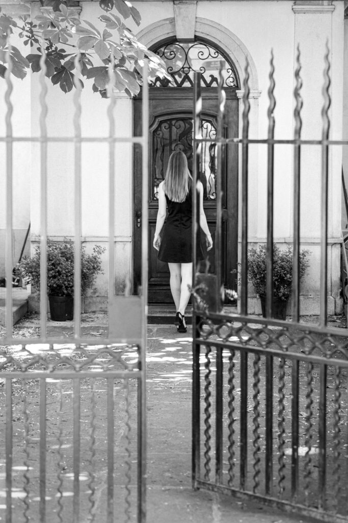 Femme en robe noire vue de dos traversant une cour vers une porte ancienne, aperçue à travers les barreaux d'une grille en fer forgé, Vieille-Ville de Genève, noir et blanc