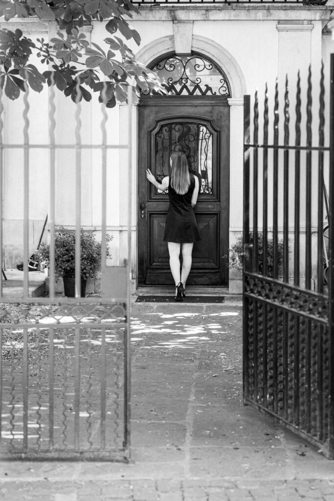 Femme en robe noire de dos devant une porte de bois cintrée, la main posée sur le battant, encadrée par une grille en fer forgé ouverte, Vieille-Ville de Genève, noir et blanc