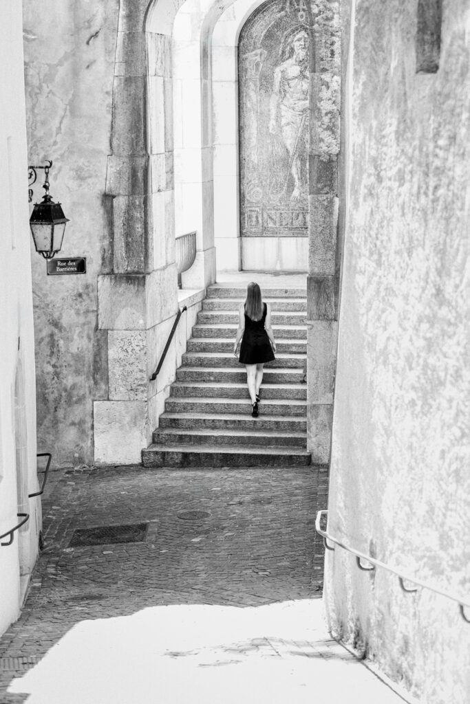 Silhouette de femme en robe noire vue de dos gravissant l'escalier du passage des Barrières vers la mosaïque Neptune de Marcel Poncet, Vieille-Ville de Genève, noir et blanc.