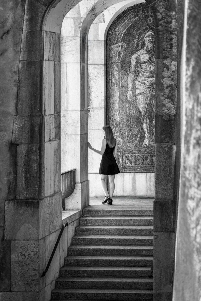 Femme en robe noire de dos, arrêtée en haut de l'escalier, la main posée sur la pierre, devant la mosaïque Neptune de Marcel Poncet, passage des Barrières, Genève, noir et blanc.