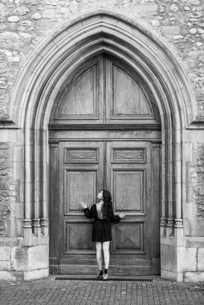 Femme en robe noire au dos ouvert, de dos et le visage levé, appuyée contre le grand portail de bois en arc brisé de l'église Saint-Germain, Vieille-Ville de Genève, noir et blanc