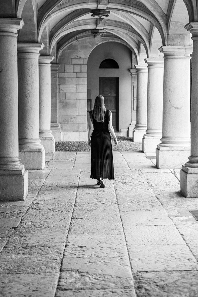 Femme en robe longue noire vue de dos marchant sous une galerie à colonnes et voûtes d'arêtes vers une porte au fond, cour de l'Hôtel-de-Ville, Genève, noir et blanc