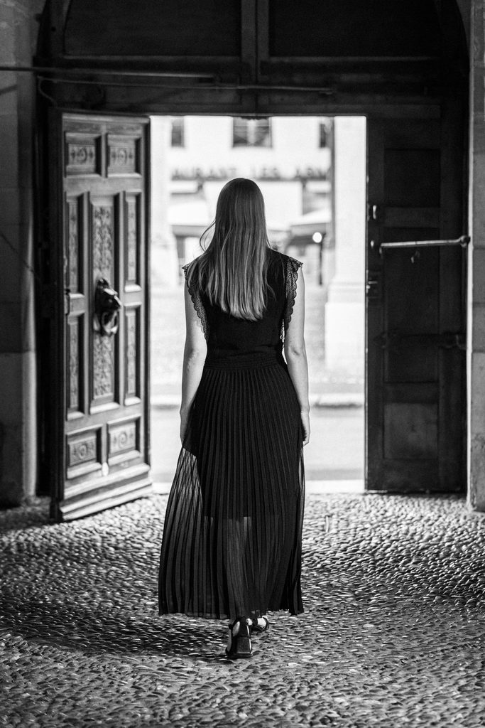 Femme en robe longue noire vue de dos, centrée dans l'ouverture lumineuse d'un porche sombre, cour de l'Hôtel-de-Ville, Vieille-Ville de Genève, noir et blanc