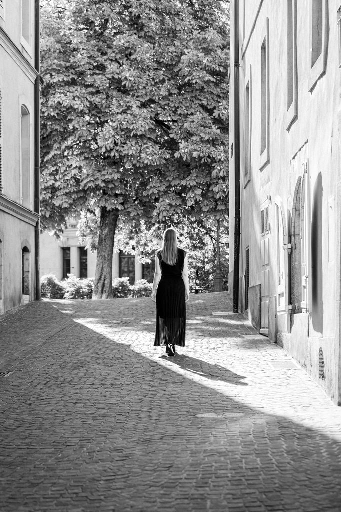 Femme en robe longue noire vue de dos marchant sur les pavés d'une rue en pente vers un grand marronnier, rue Maurice, Vieille-Ville de Genève, noir et blanc