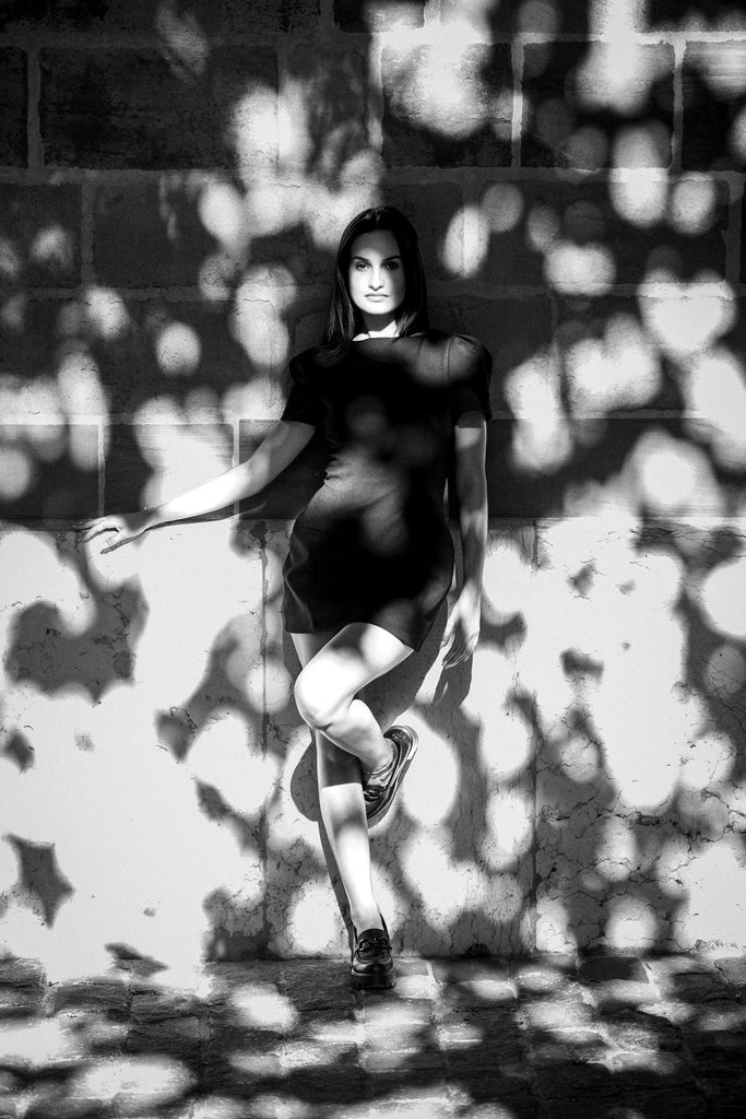 Femme en robe noire adossée au mur de la cathédrale Saint-Pierre, le corps traversé par les ombres portées du feuillage d'un arbre, Vieille-Ville de Genève, noir et blanc
