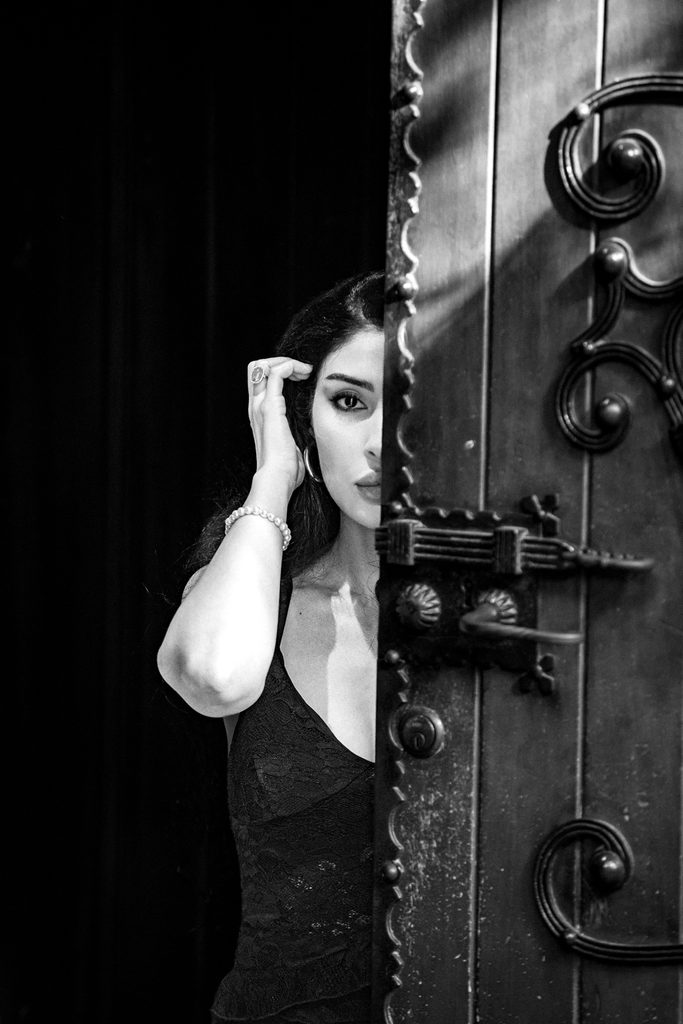 Femme en robe noire à demi sortie d'une porte entrouverte, la main aux cheveux, la lumière découpant son visage, chapelle des Macchabées, Genève, noir et blanc